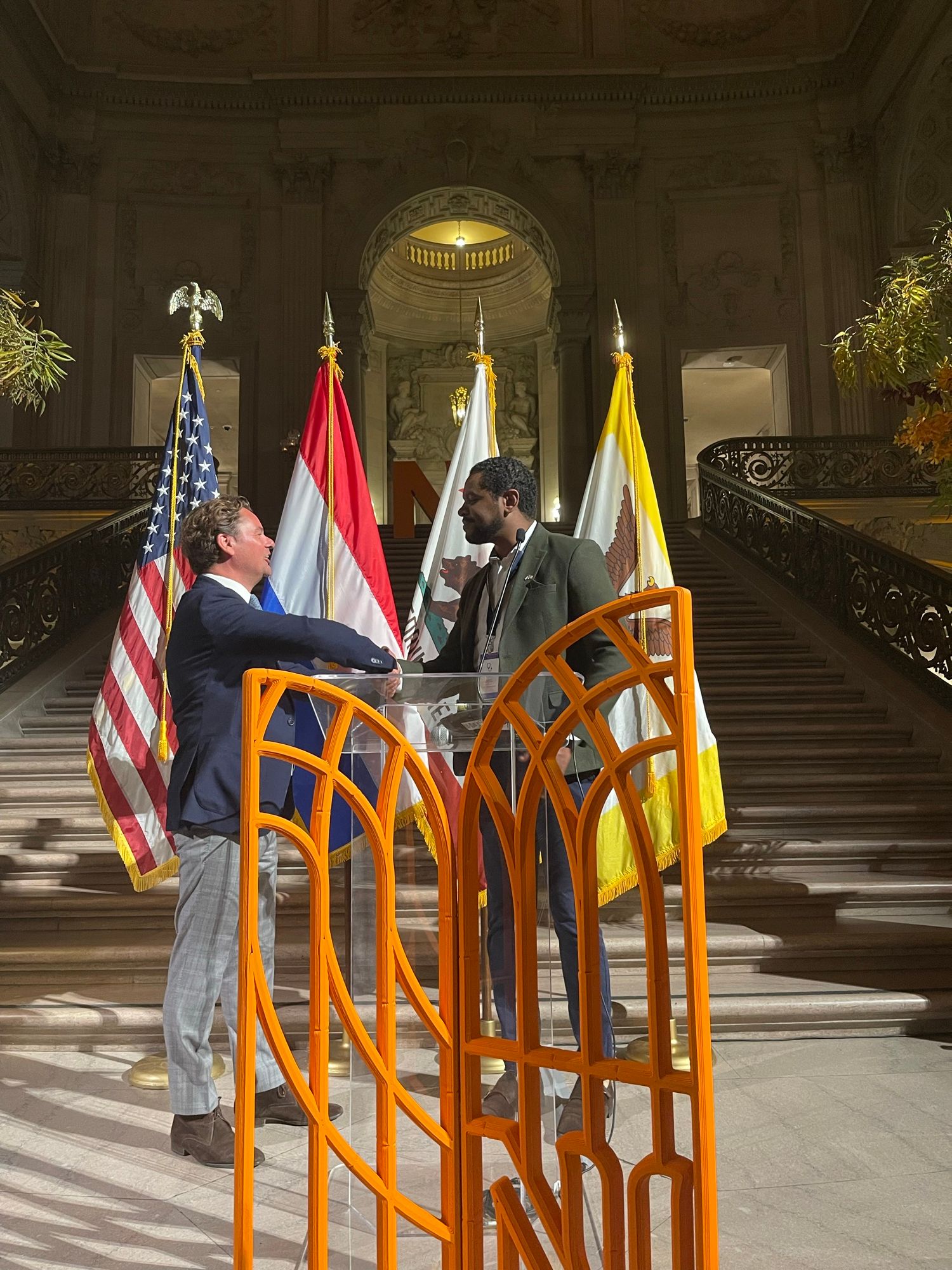 Michel Tzsfaldet shaking hands during the Netherlands economic mission to California, at San Francisco City Hall beside United States and Dutch flags.