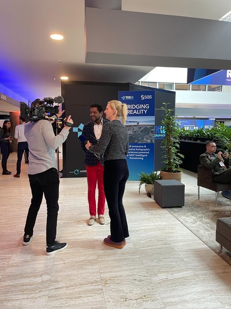 Michel Tzsfaldet being interviewed on camera at the REAIM 2023 summit, standing in front of a Tekle Holographics and SAS banner.